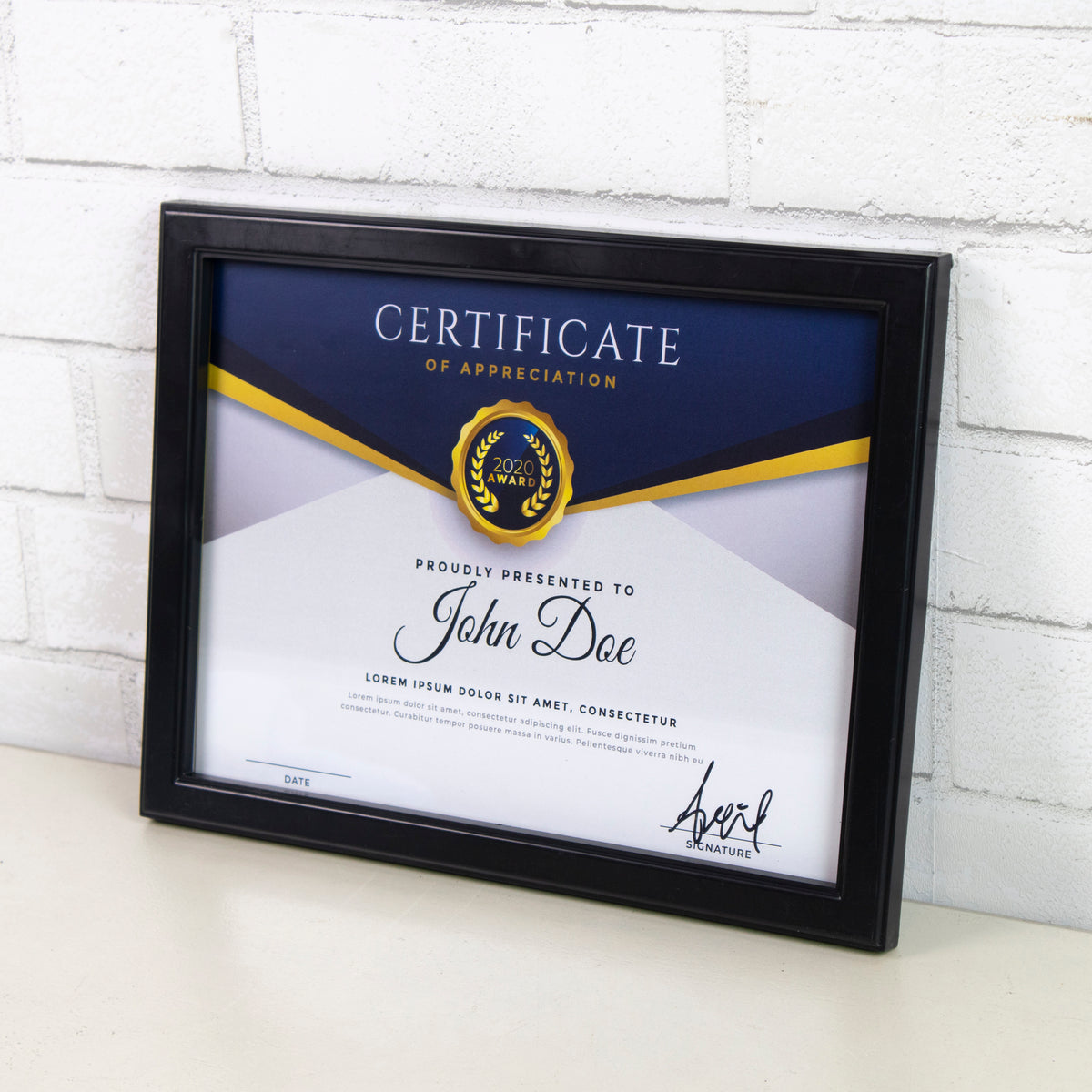 Black frame displaying a certificate of appreciation horizontally on a tabletop against a white brick wall.
