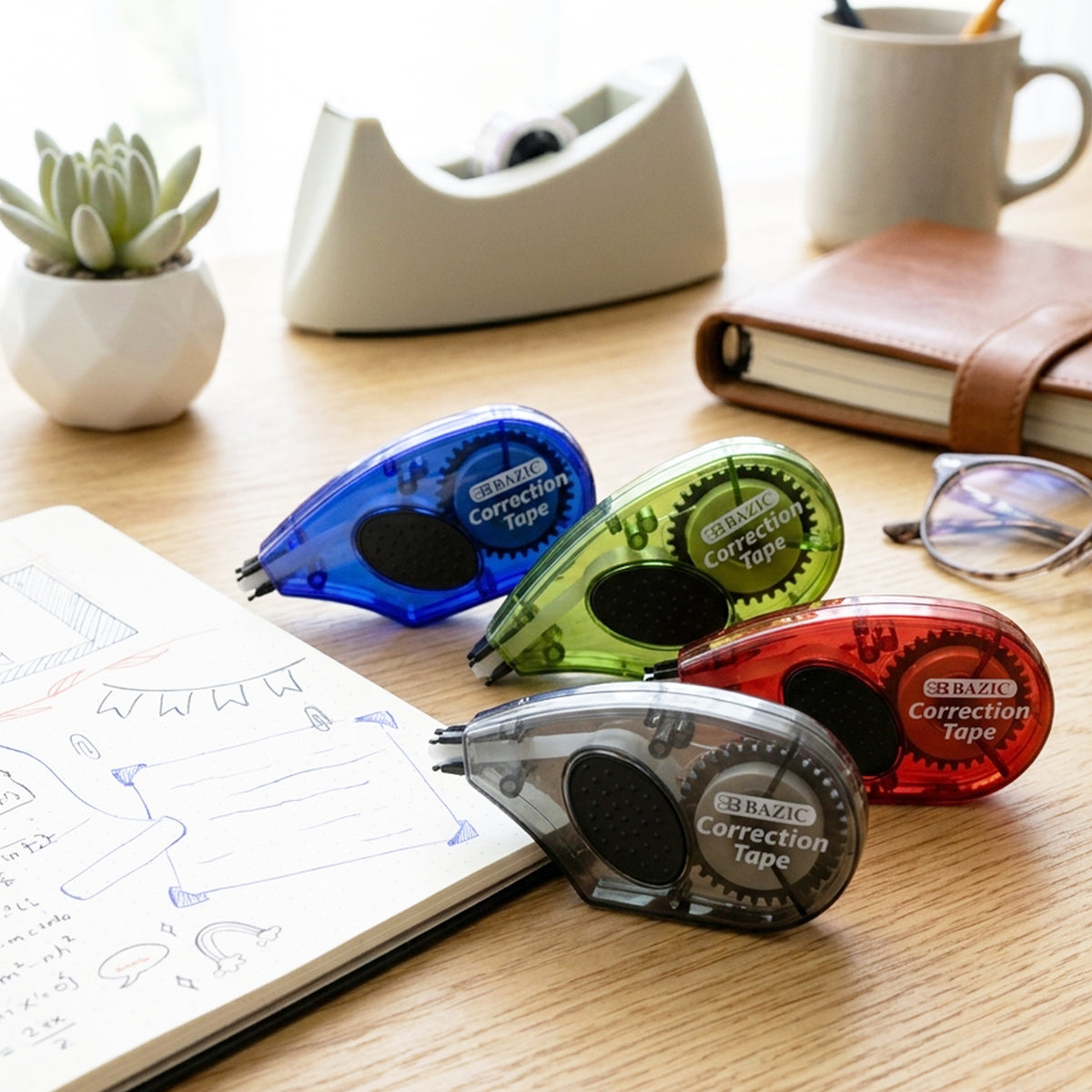 Four correction tape dispensers with translucent blue, green, red and gray cases and black grips on a desk beside a notebook.
