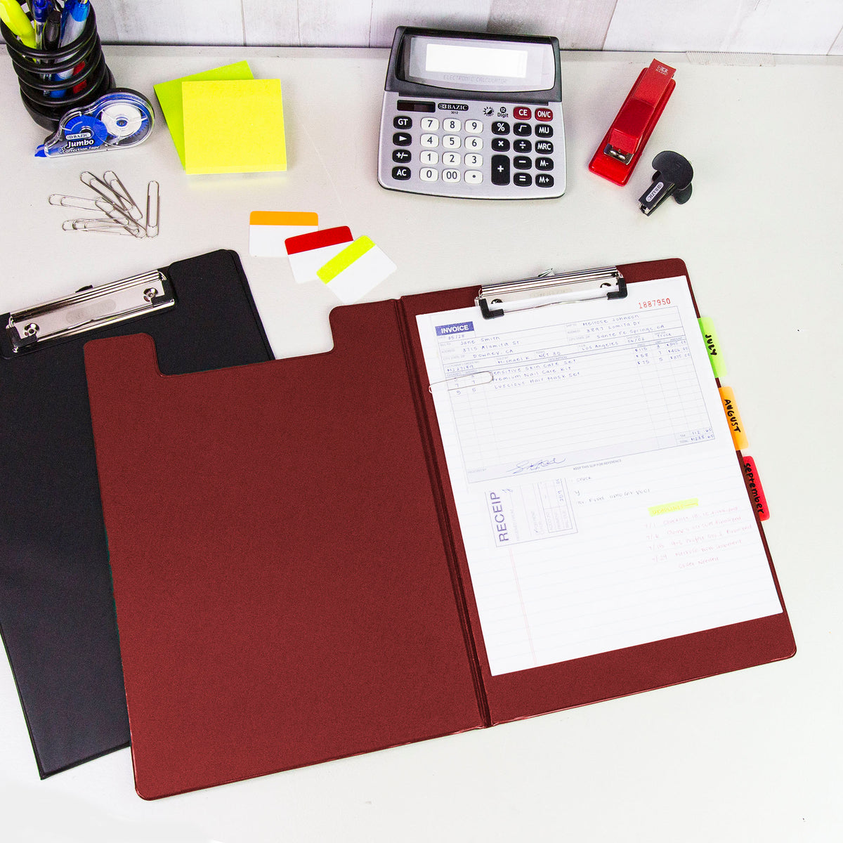 Open burgundy clip folder holds paperwork with month tabs on a desk beside a black folder and office supplies.