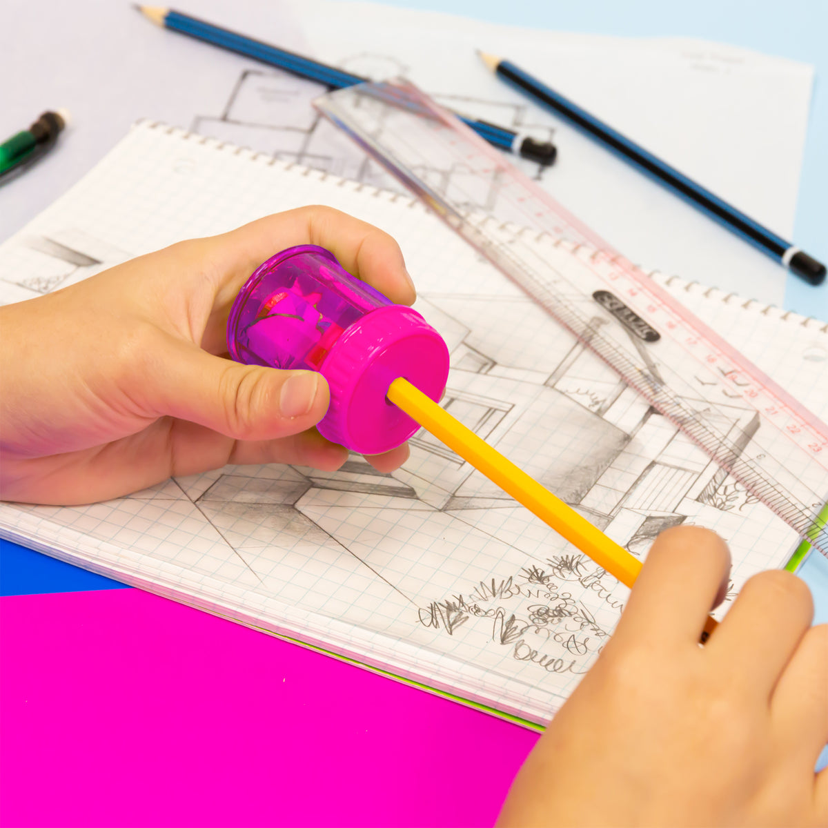A person sharpens a yellow pencil with a translucent pink sharpener over a notebook of sketches.