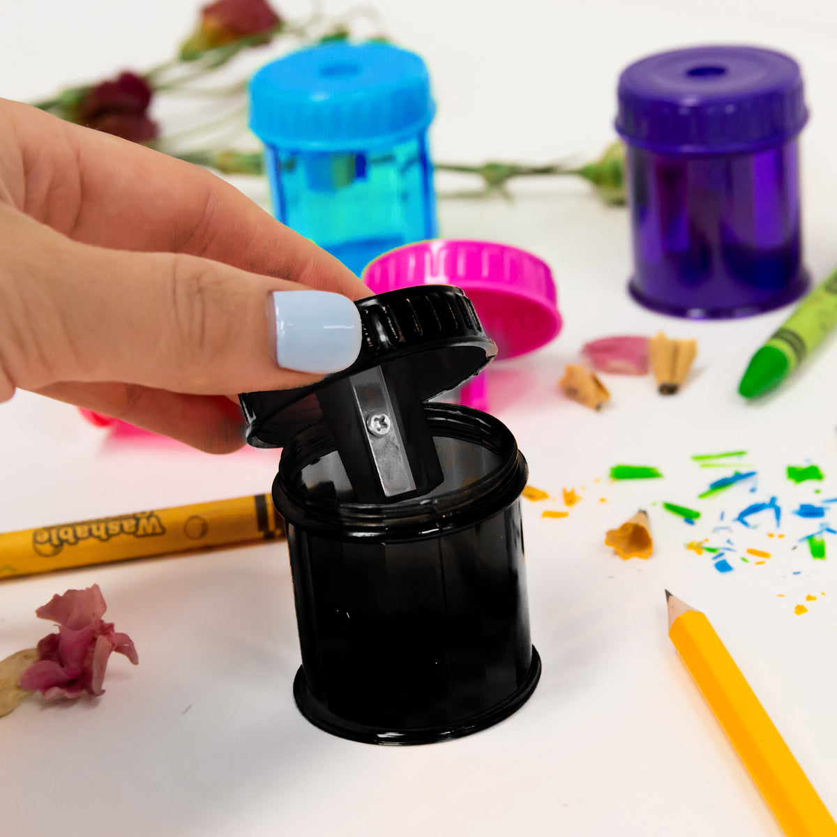 A hand lifts the blade lid from a black sharpener's shavings container, with blue, pink and purple sharpeners nearby.