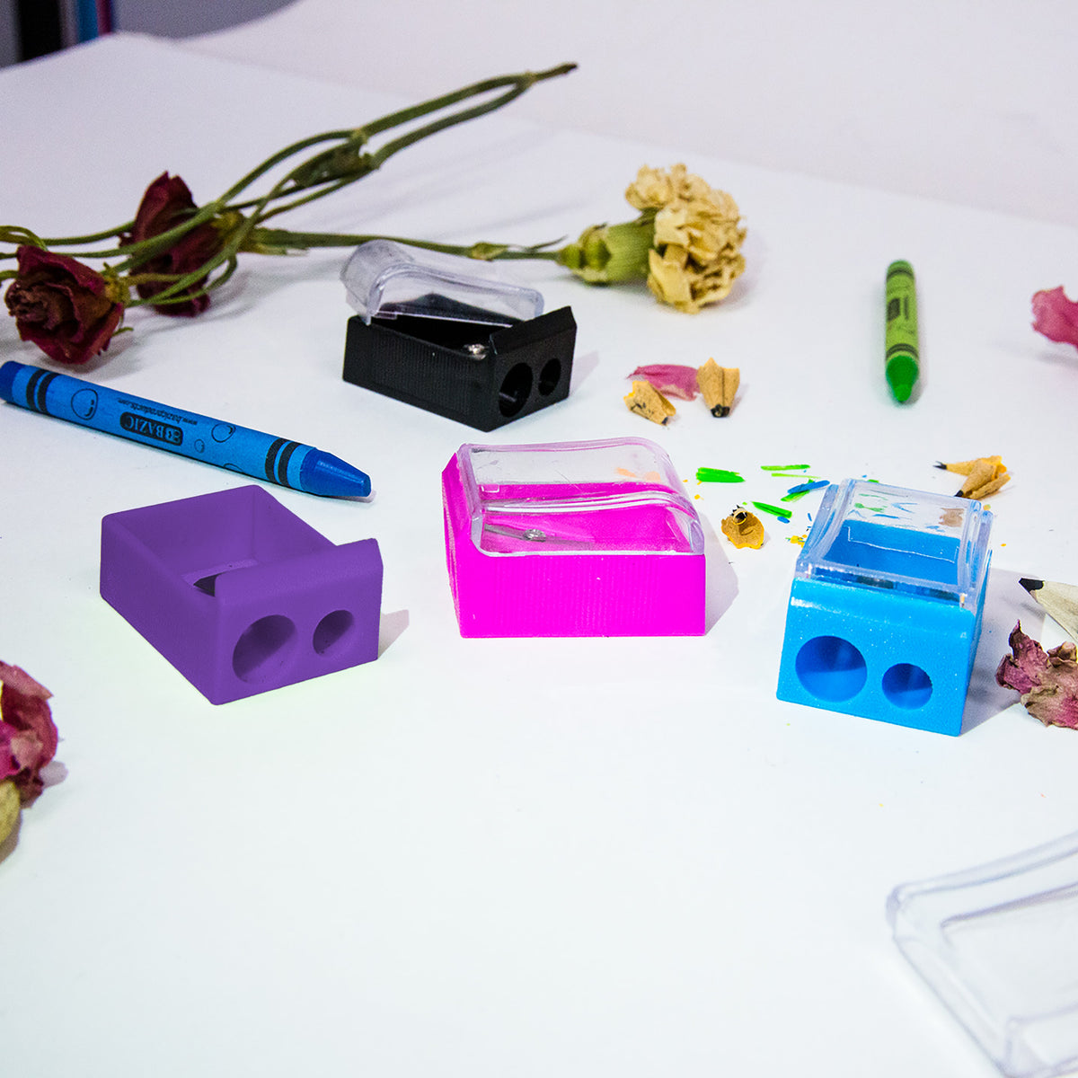 Four sharpeners in black, purple, pink and blue with two hole sizes and clear lids, beside crayons and shavings.