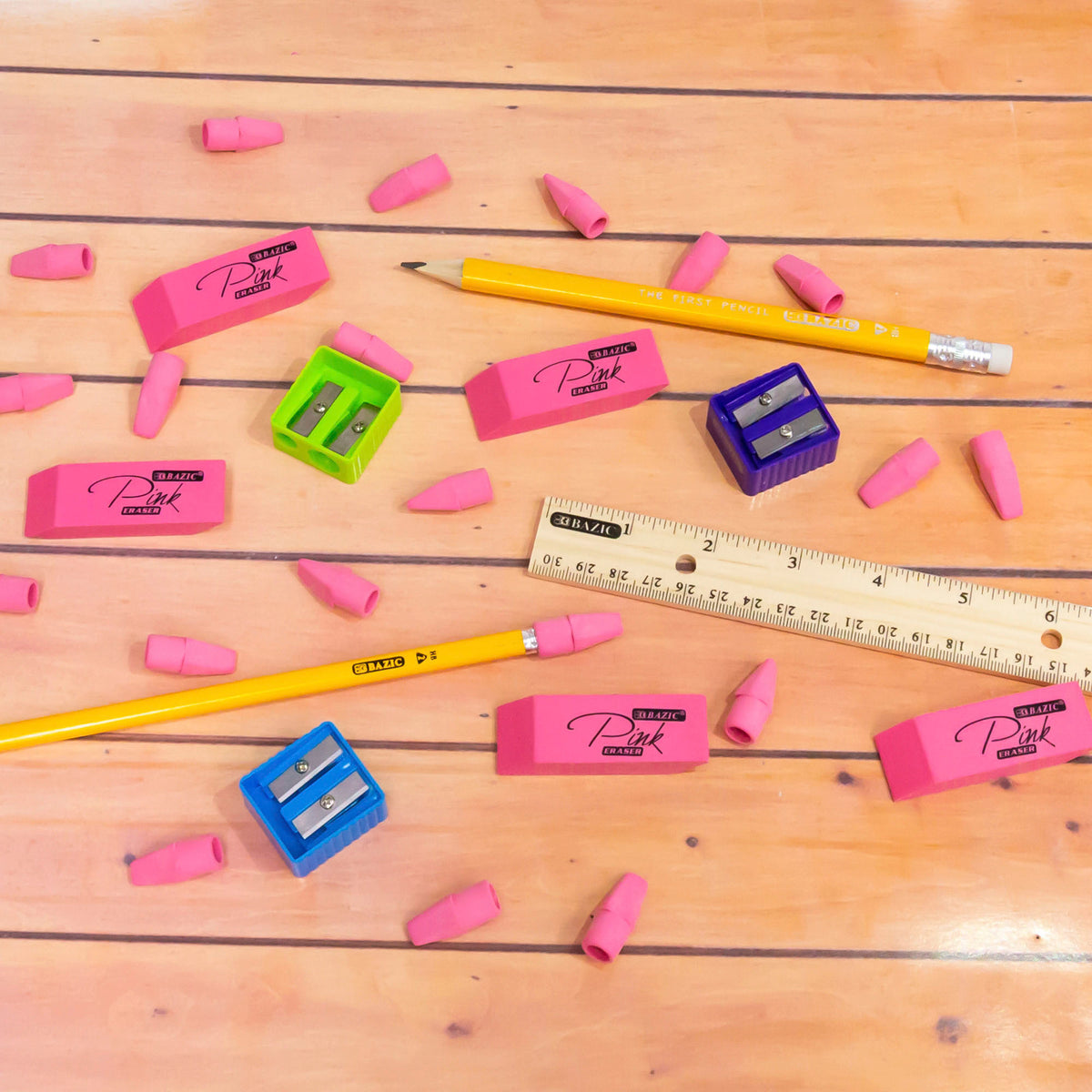 Pink eraser caps scattered on a wooden surface with pencils, block erasers, colorful sharpeners and a ruler.