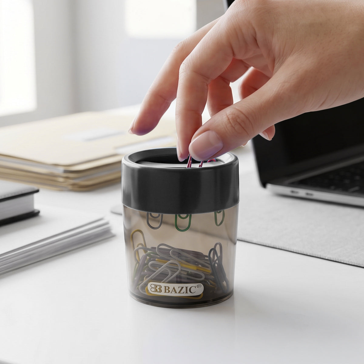 A person picks a colored paper clip from a clear holder with a black magnetic top on a desk beside a laptop.