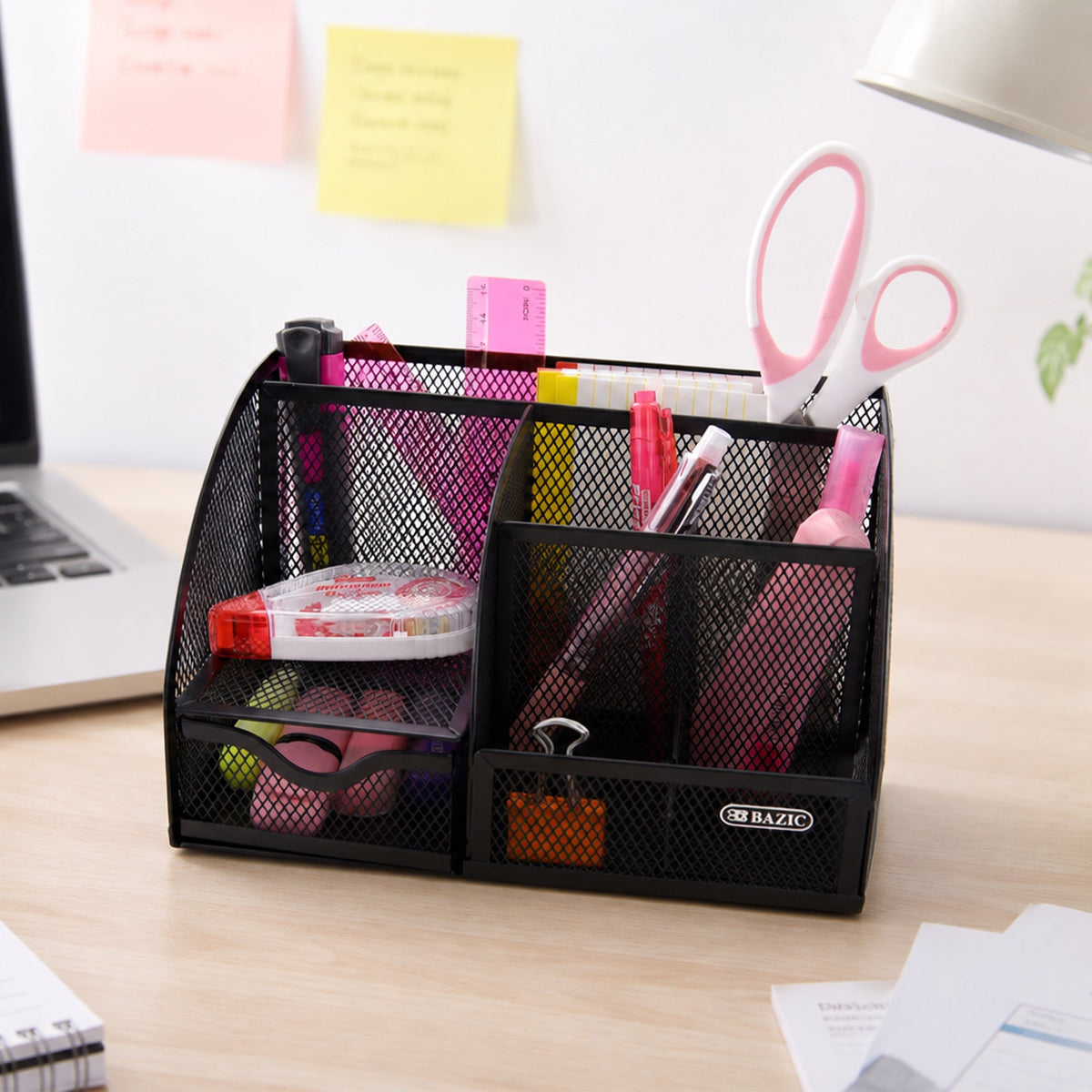 Black mesh organizer on a desk holds scissors, pens, a ruler and binder clips in divided compartments and a drawer.