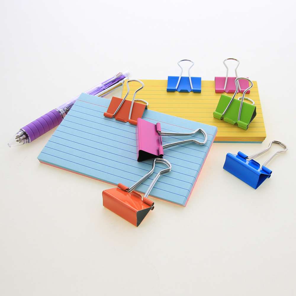 Seven blue, pink, orange and green binder clips with silver wire handles on and beside lined index cards and a purple pen.