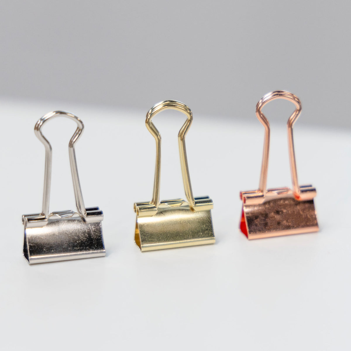 Three shiny binder clips in silver, gold and rose gold, with wire handles raised.