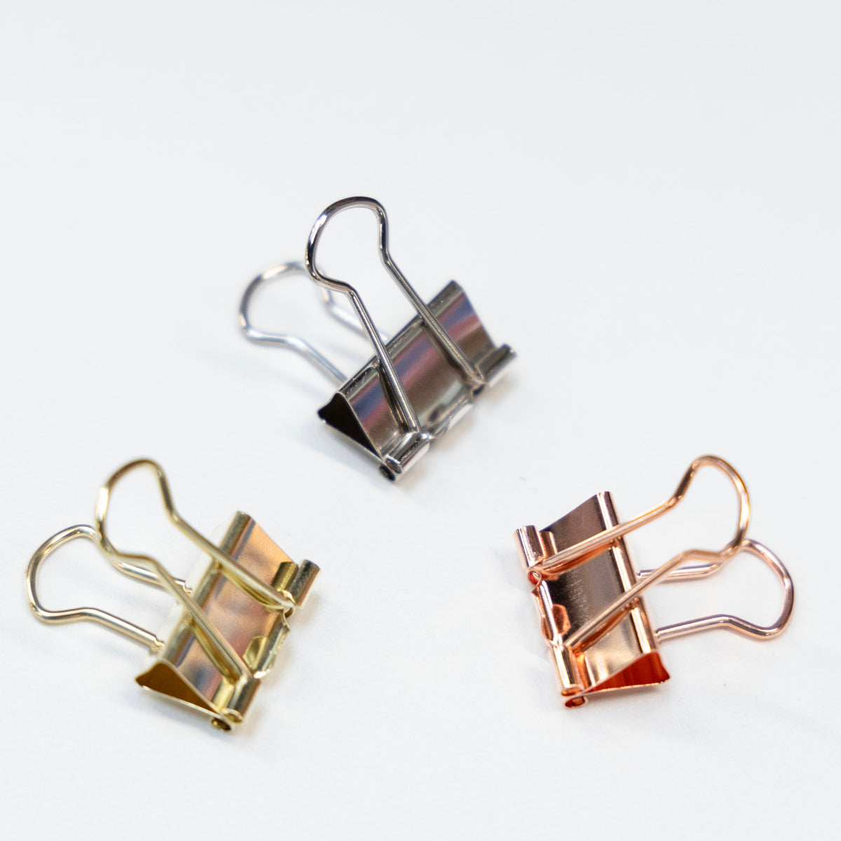 Three shiny binder clips in silver, gold and rose gold, with raised wire handles on a white surface.
