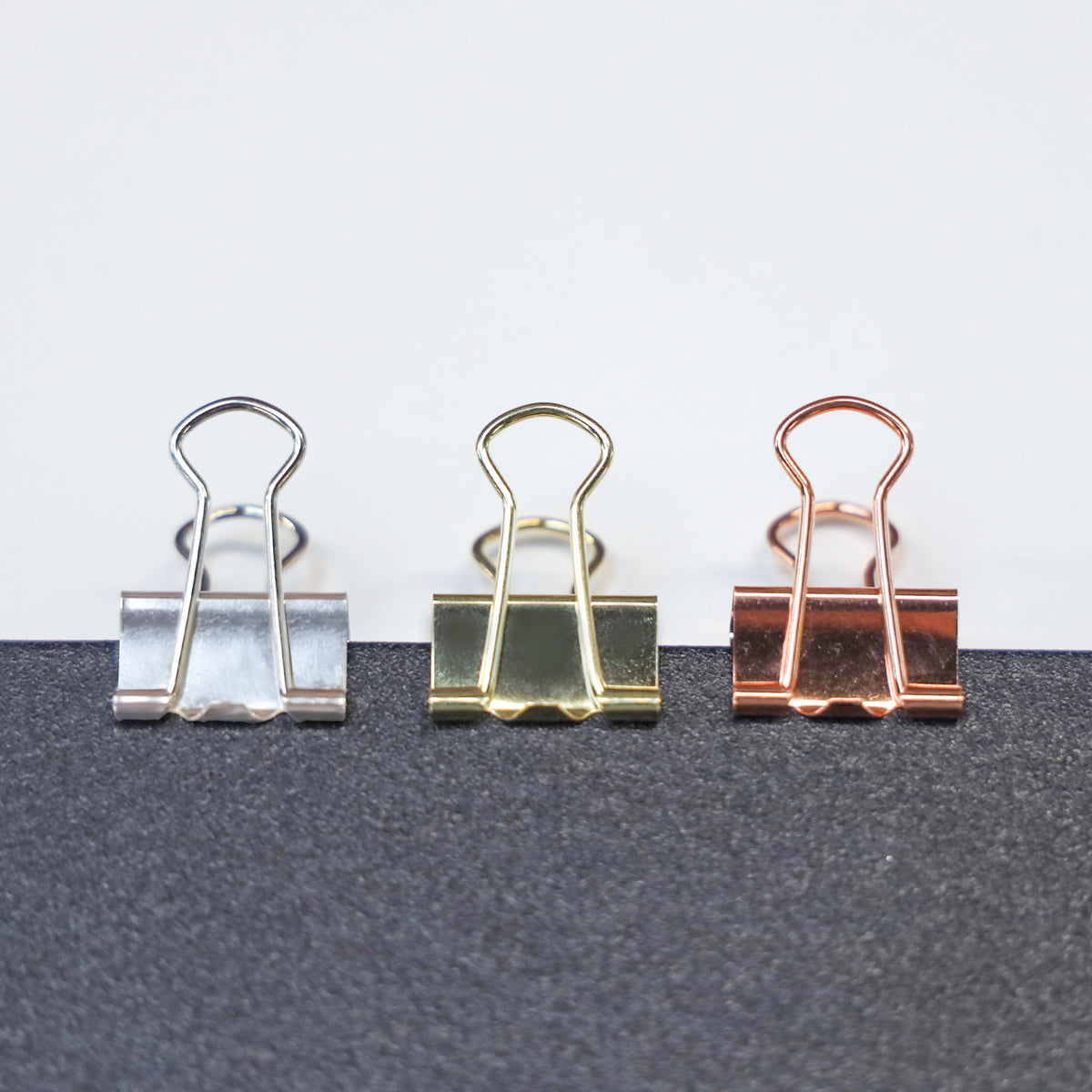 Three binder clips in silver, gold and copper finishes, lined up with wire handles raised on a dark surface.