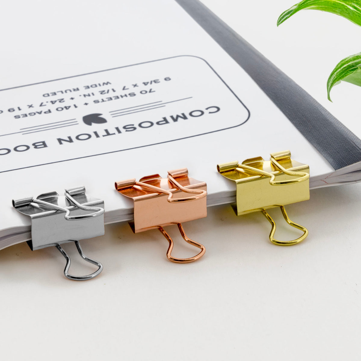 Three metallic binder clips in silver, rose gold and gold grip the edge of a composition notebook.