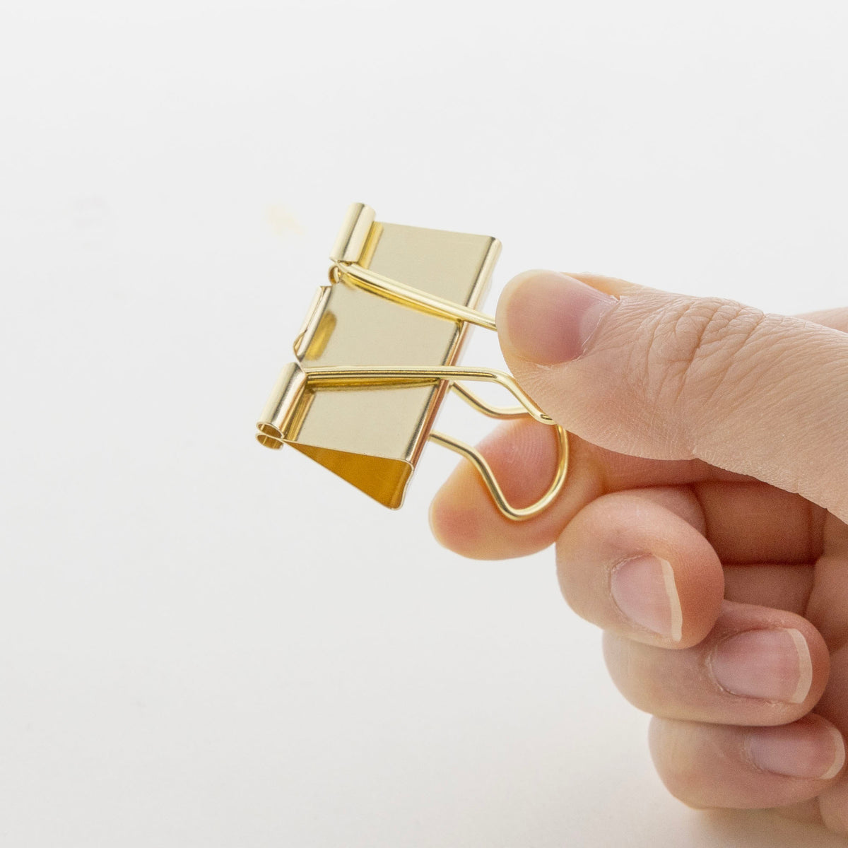 A hand squeezes the wire handles of a gold metallic binder clip to open its jaws.
