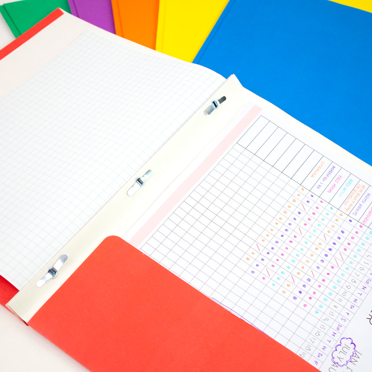 Open red folder with three metal prongs holding graph paper and a pocket holding a chart; assorted colors behind it.
