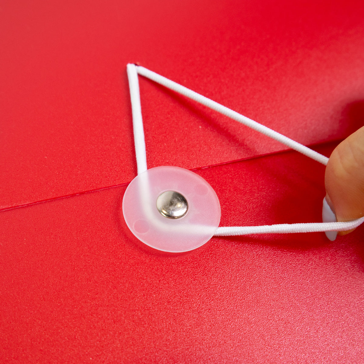Close-up of a hand pulling the red folder’s white elastic cord around a translucent button closure.