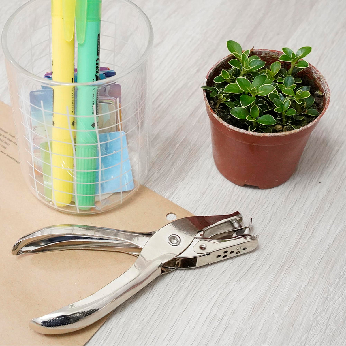 Silver metal hole punch rests on punched kraft paper beside a pen cup and potted plant.