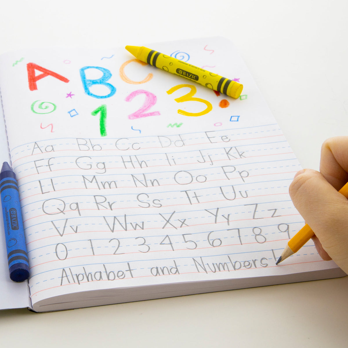 A hand uses a pencil to practice letters and numbers on primary-ruled lines below colorful crayon drawings.