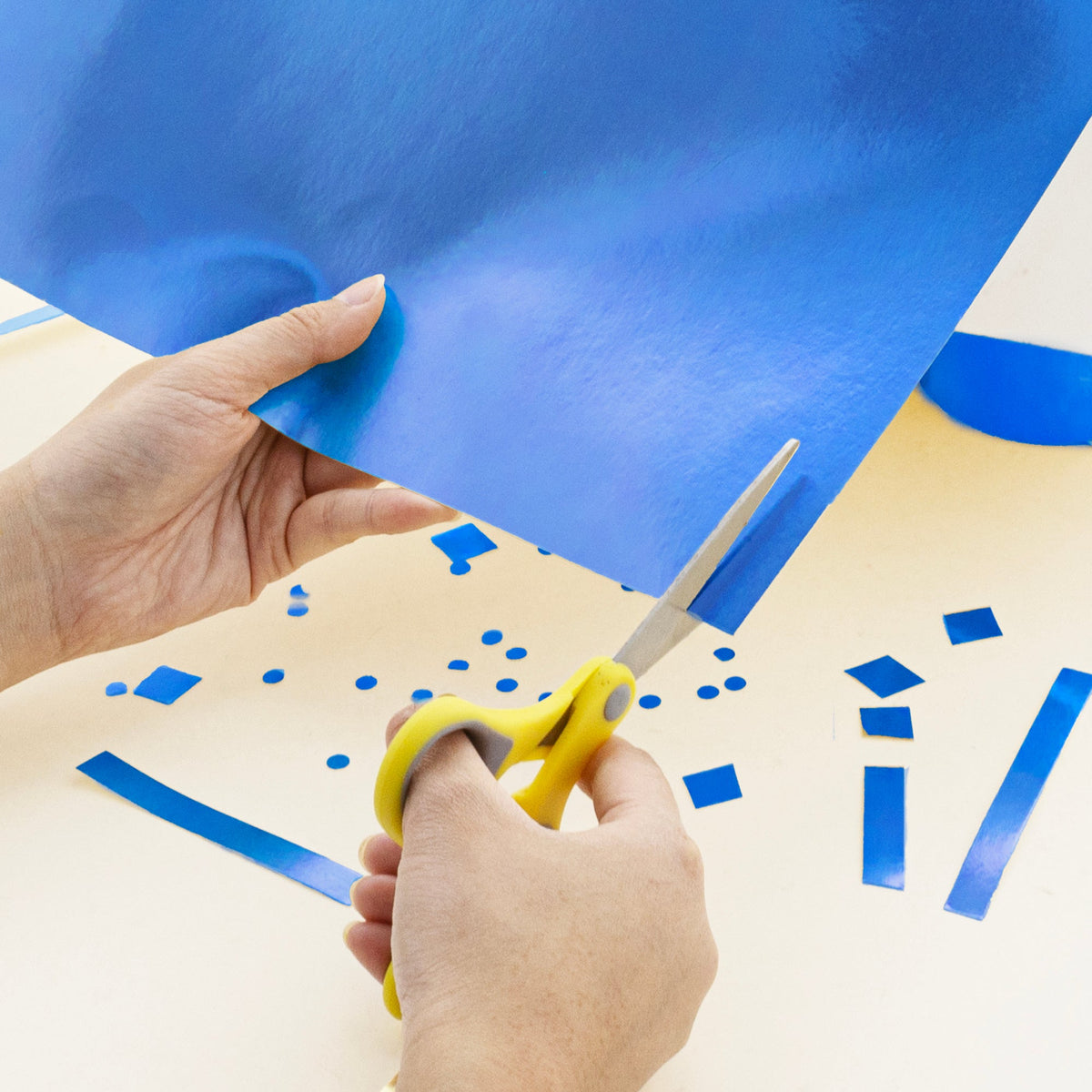 A person cuts shiny blue poster board with yellow-handled scissors, with blue scraps scattered on the table.