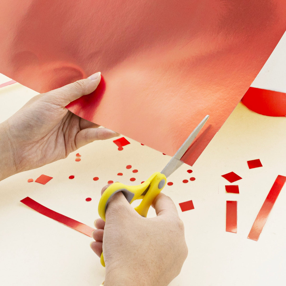 A person cuts shiny red poster board with yellow-handled scissors, with red cutouts scattered on the table.