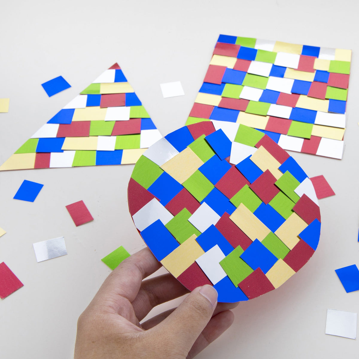 A hand holds a circular mosaic craft with colorful rectangles, beside matching triangle and square crafts.