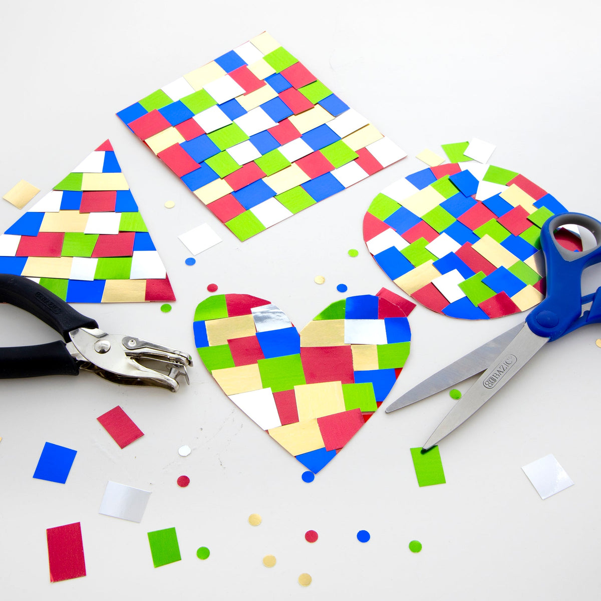 Colorful woven paper crafts in heart, circle, triangle and square shapes, with scissors and a hole punch on a table.