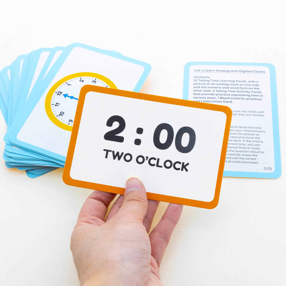 A hand holds an orange-bordered card reading 2:00, two o’clock, beside blue-bordered clock cards and instructions.