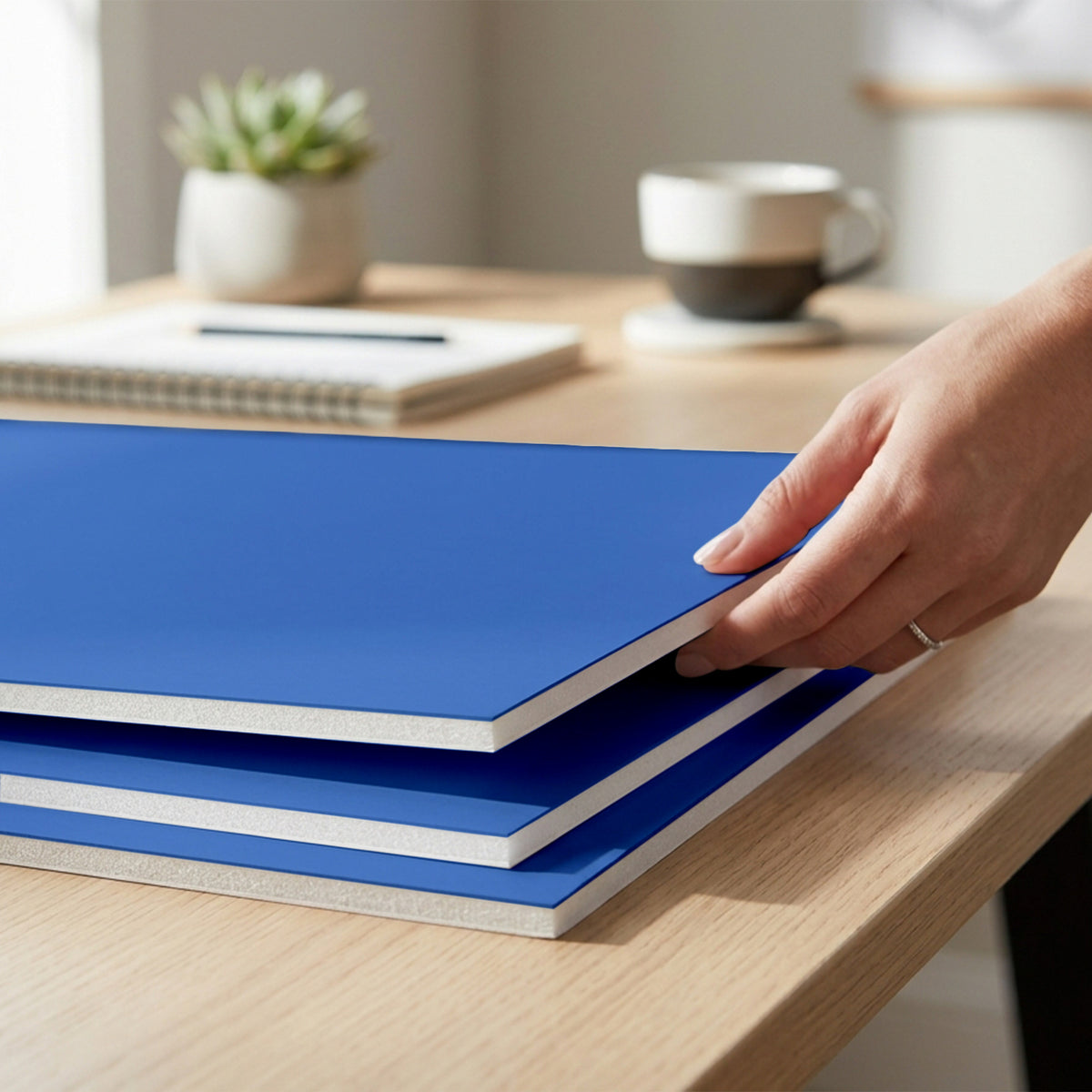 A person lifts the top of three stacked blue foam boards on a wooden desk, showing their white foam edges.