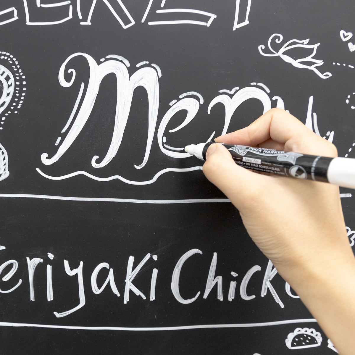 A person uses a white chalk marker to write decorative menu lettering on the black chalkboard.