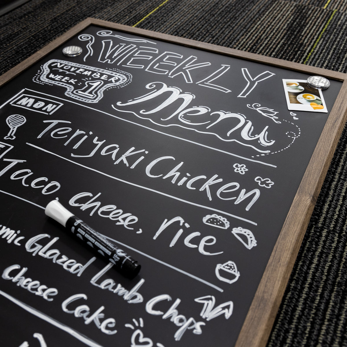 Wood-framed black chalkboard with a white weekly menu, two round magnets, a small photo and a chalk marker.