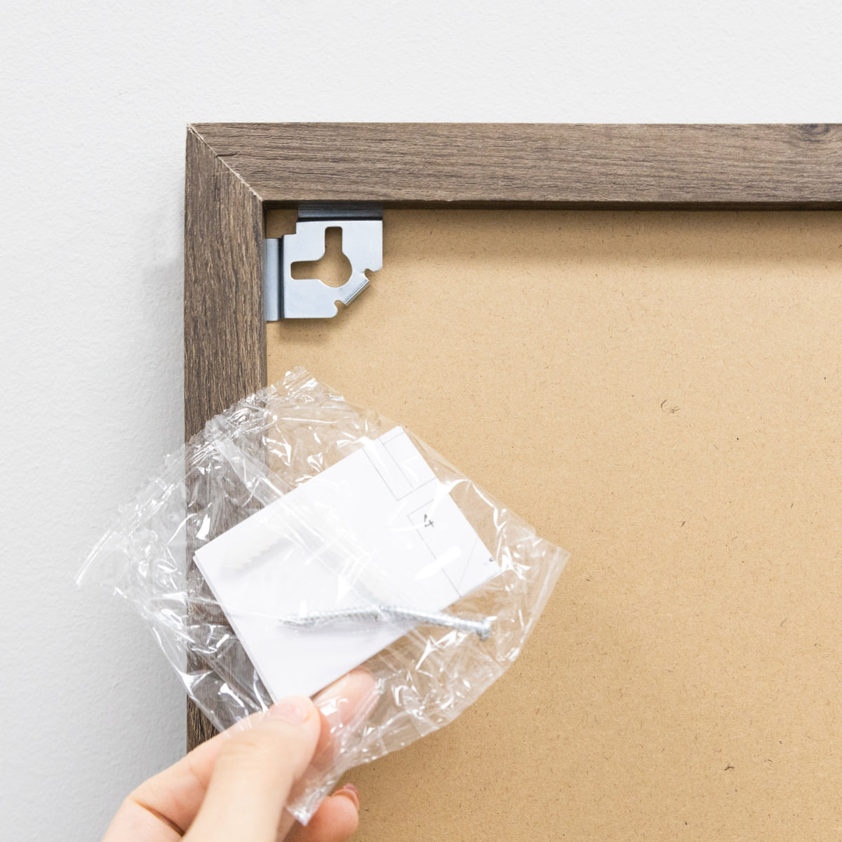 Back corner of the wood frame with a metal hanging bracket and a hand holding a bag of mounting hardware.