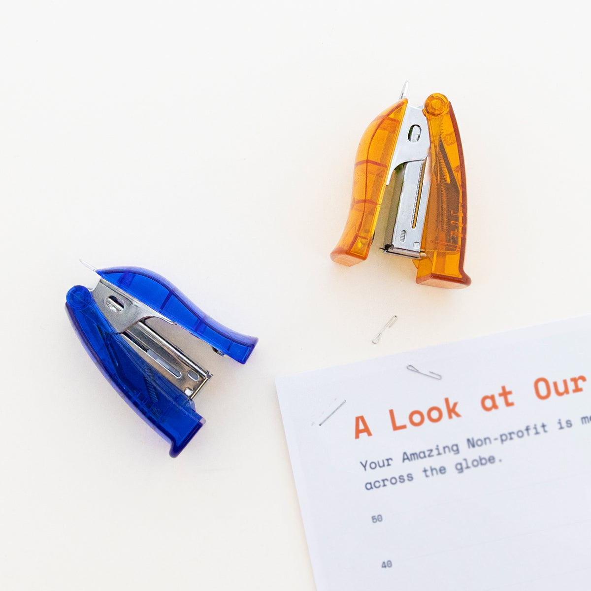 Blue and orange translucent mini staplers shown open beside loose staples and a stapled sheet of paper.