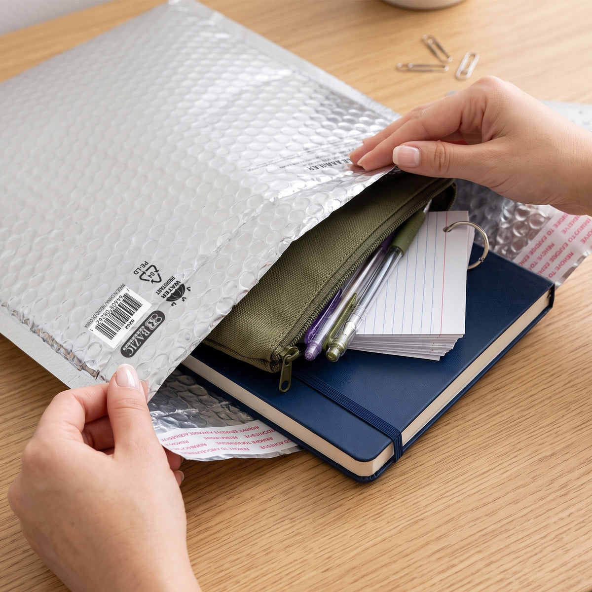 Hands hold open a silver bubble mailer containing a notebook, pencil pouch, pens and ring-bound index cards.