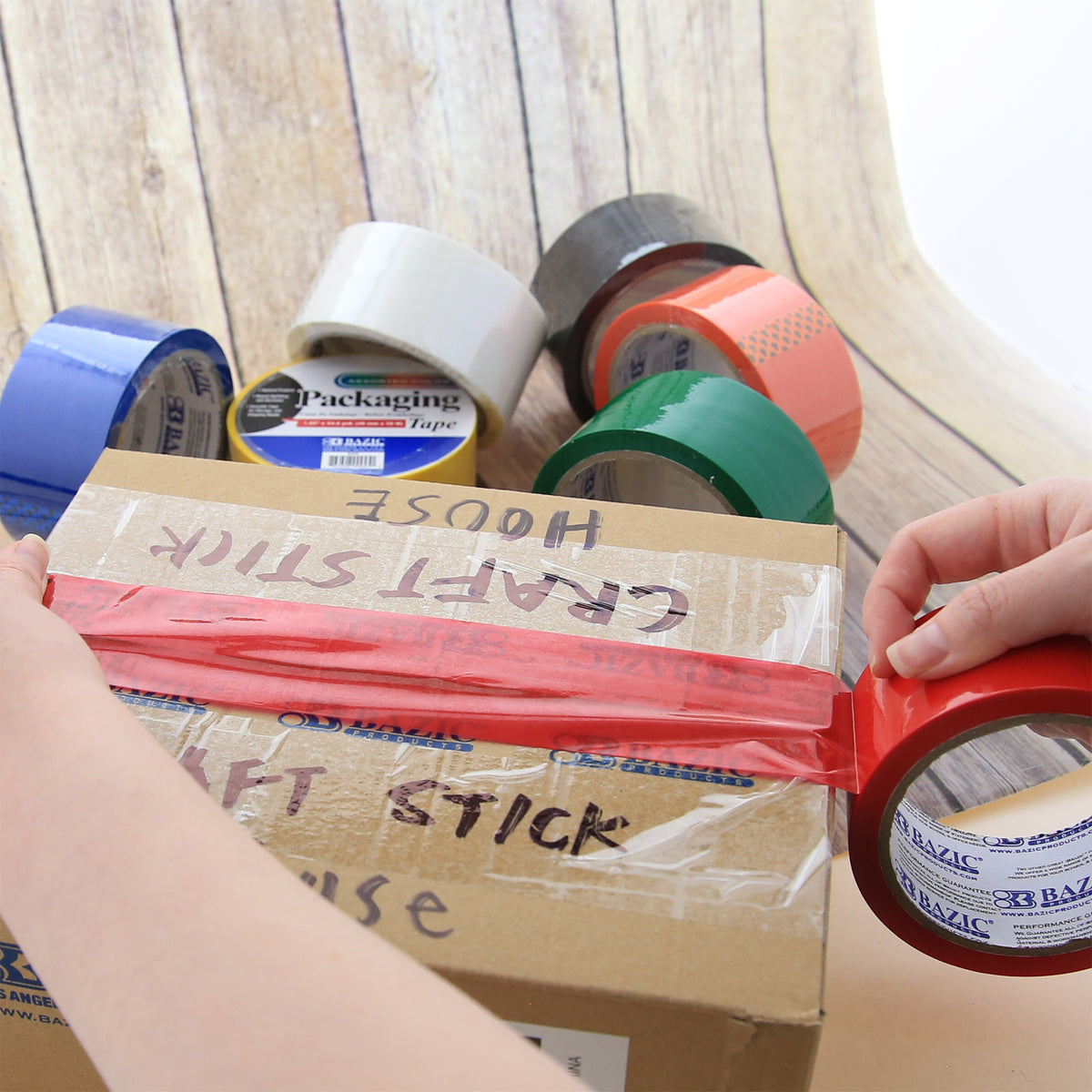 A person seals a cardboard box with red packaging tape, with assorted colored rolls nearby.
