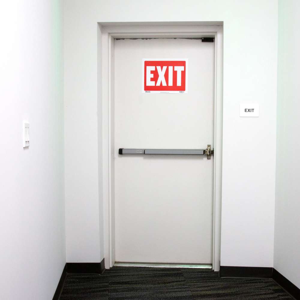 Red sign with large white EXIT lettering mounted on a white door above a push bar.