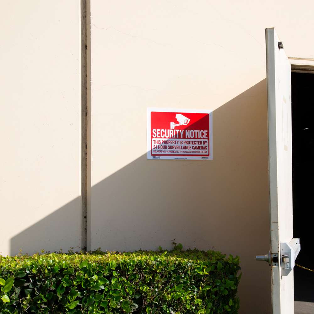 Red security notice sign beside an open door reads: This property is protected by 24 hour surveillance cameras.
